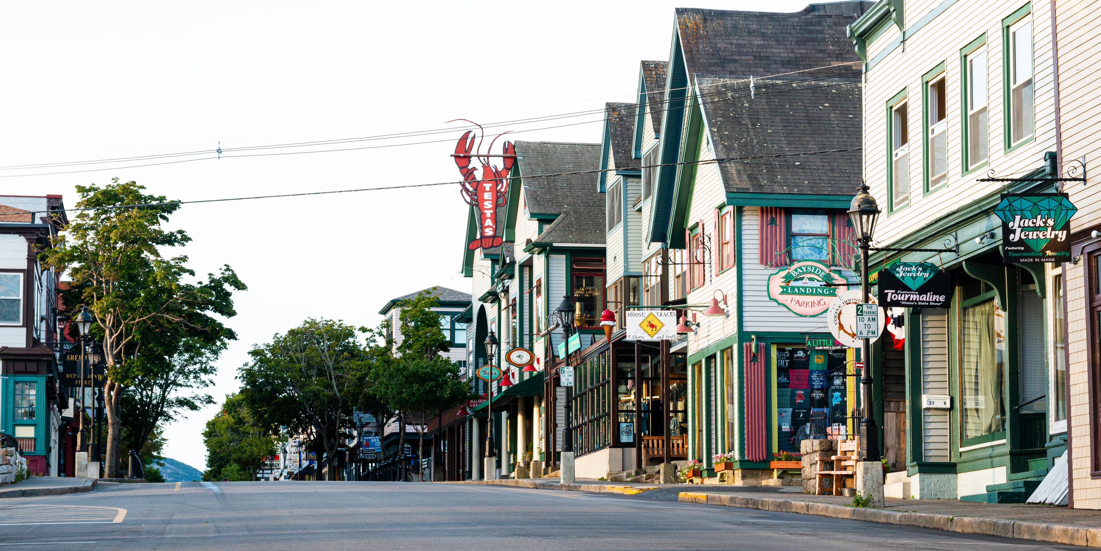 Bar Harbor village waterfront in early May, empty Main Street, window boxes being filled with flowers, Acadia mountains rising blue in the background across the harbor, morning fog lifting