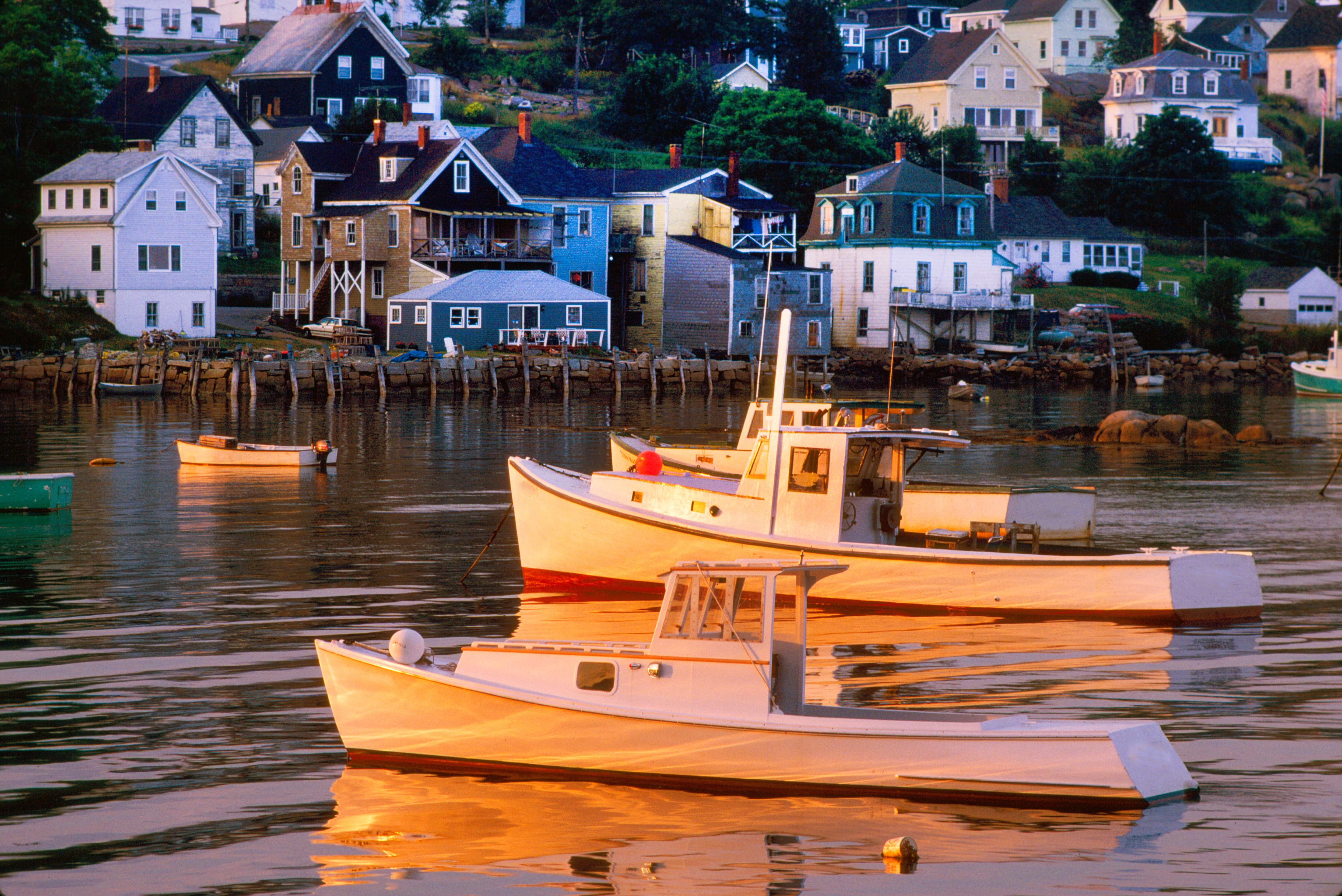 Stonington harbor at first light, lobster boats heading out to sea, wooden-shingled fish houses on stilts over grey water, an old granite quarry visible on the far shore, mist on the islands